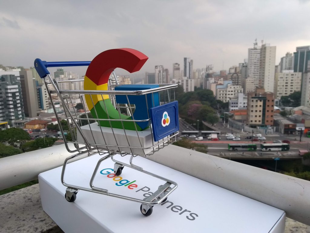 foto na varanda do prédio da Goobec. Ao fundo está a avenida 23 de Maio com vários carros e ônibus transitando. Em destaque na foto, há um pequeno carrinho de supermercado de 30 cm. Dentro dele há uma mini escultura do G do Google. Esse G do Google mede 15 centímetro e ele é colorido, nas cores: azul, amarelo, vermelho e azul.